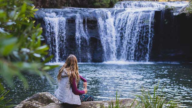 A woman in a jacket sits in front of a waterfall
