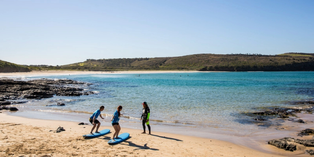 A surf instructor in a wetsuit stands in front of two people on surfboards on a sunny, sandy beach framed by green hilled shores.